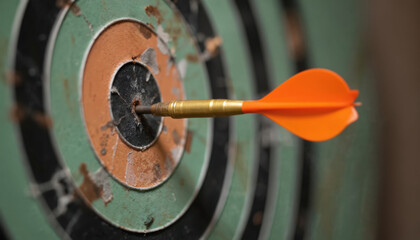 Dart hits bullseye on worn dartboard. Orange dart fletching is clearly visible against green and black concentric circles. Focus on precision and achieving goals in sports, business, or life.