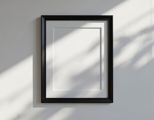 Rectangular black picture frame hangs on clean white wall. Empty blank matting inside frame is ready for custom artwork or photo placement. Natural light casts shadows across the surface.