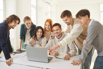 Friendly group of young people gathering around table, looking at laptop screen and smiling....