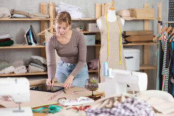 Careful young woman tailor recording dress parameters using tablet in sewing studio