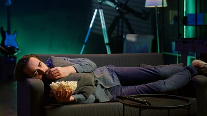 Depressed man at home lounged on couch feeling low, seeking comfort by watching movies on TV and eating popcorn. Glum person laying down, suffering from depression, watching TV to feel better, camera