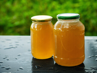 Two glass jars of golden bee honey are on a wet table, green grass field and sky in the background....