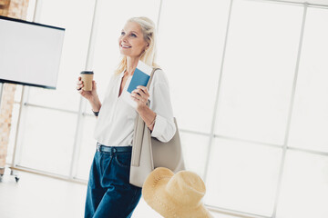 Mature woman holding a cup and documents in an airport terminal, excitedly anticipating her...