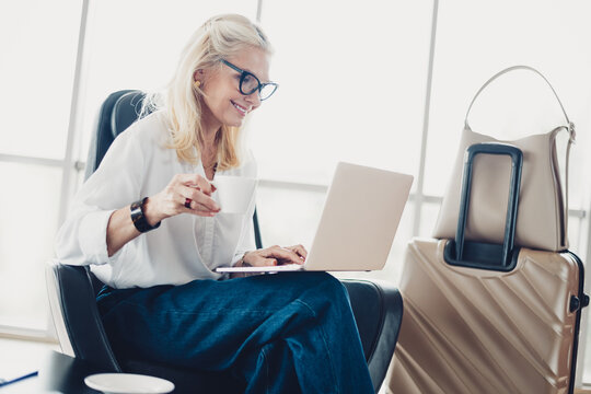 Cheerful woman sitting in an airport lounge, enjoying a cup of coffee while working on a laptop with luggage beside her