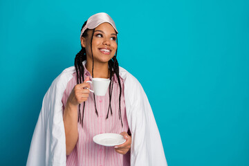 Young woman in sleepwear enjoying a cup of tea against a vibrant blue background, embracing a...