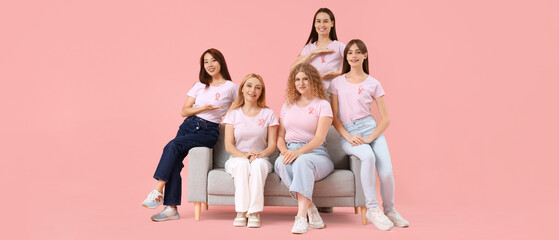 Beautiful women with pink ribbons sitting on sofa against color background. Breast cancer awareness...