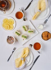 Tea setting for two with white tableware and yellow napkins, cucumber sandwiches and oatmeal cookies on a light tablecloth, top view.