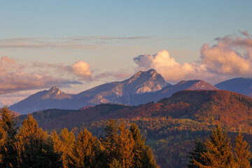 Mountainous landscape at dawn with warm sunlight illuminating forests and clouds, creating a...