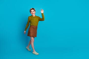 Happy young woman with short hair wearing a khaki shirt and brown skirt waves her hand against a...