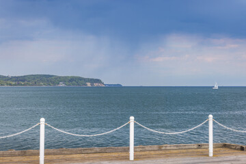 The coast of the Baltic Sea in northern Poland. A serene seascape featuring calm blue waters, a distant sailboat, and a lush green shoreline, framed by a wooden dock and white posts.