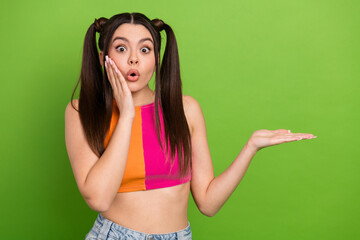 Surprised young woman with ponytails presenting copy space, colorful outfit, and green background...