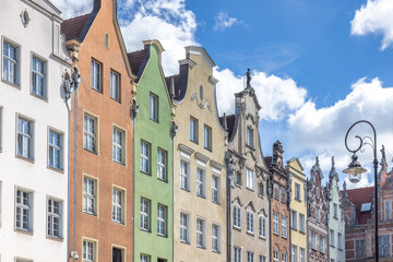 Gdansk, a city on the Baltic coast of northern Poland, Europe. Colorful historic buildings line the street, each with unique architectural details and hues against a bright blue sky with fluffy clouds