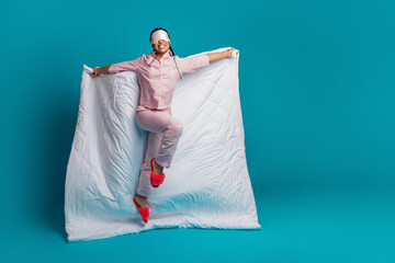 Pretty woman playfully posing in pink pajamas with a duvet and sleep mask on a vibrant blue studio...
