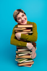Smiling young woman in casual attire holding a tall stack of book in a vibrant studio setting