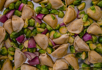 Uncooked pierogies, Brussels sprouts, and red onion on a baking sheet, ready for the oven
