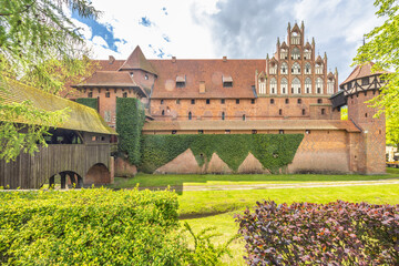 The Malbork Castle, a brick Gothic castle in northern Poland, Europe. Majestic brick castle with fortified walls and a wooden bridge, surrounded by lush greenery under a partly cloudy sky.