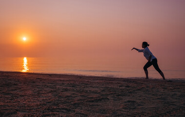 Woman practicing Tai Chi on beach at sunrise, graceful silhouette against calm sea and orange-pink...