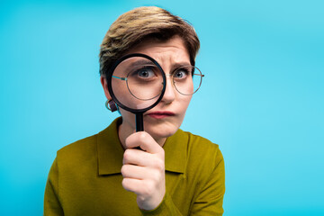 Young woman with stylish look holding a magnifying glass over a blue background expressing...
