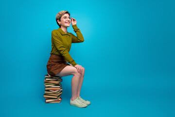 Stylish young woman wearing a khaki shirt seated on stacked book with a blue background, showcasing...