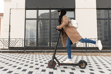 A young woman in a beige coat rides an electric scooter during the day near office buildings.