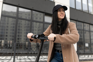 A young woman in a beige coat rides an electric scooter during the day near office buildings.