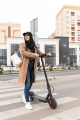 A young woman in a beige coat walks with an electric scooter through a pedestrian crossing in the...