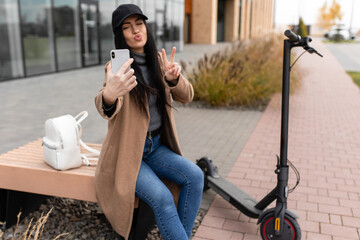Smiling young woman in a beige coat sits on a bench near an electric scooter and takes a selfie on...