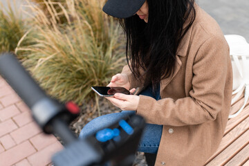 Young woman in a beige coat sits on a bench near an electric scooter and uses a smartphone against the backdrop of office buildings.