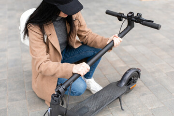 Young woman in a beige coat folds an electric scooter in the middle of a city street. A person uses an environmentally friendly form of transport in the form of an electric scooter.
