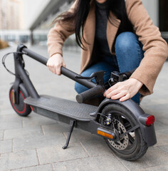 Young woman in a beige coat folds an electric scooter in the middle of a city street. A person uses an environmentally friendly form of transport in the form of an electric scooter.