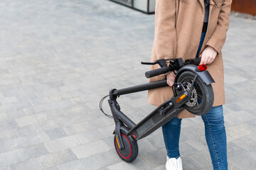 Young woman in a beige coat carries an electric scooter in his hands on the street. A person uses an environmentally friendly form of transport.