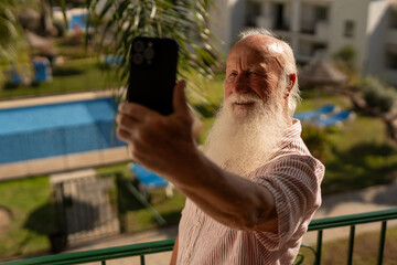 Happy senior man with long white beard taking selfie with smartphone on balcony, enjoying sunny vacation in tropical resort