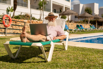 Senior freelancer enjoying remote work by the pool, relaxed elderly businessman using laptop...