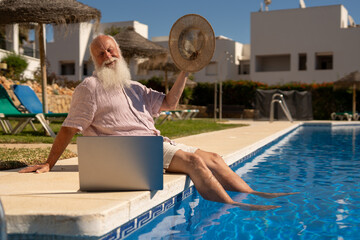 Elderly man enjoying retirement life in Spain, relaxing by the pool with feet in the water and laptop nearby, symbol of digital freedom and peaceful senior lifestyle