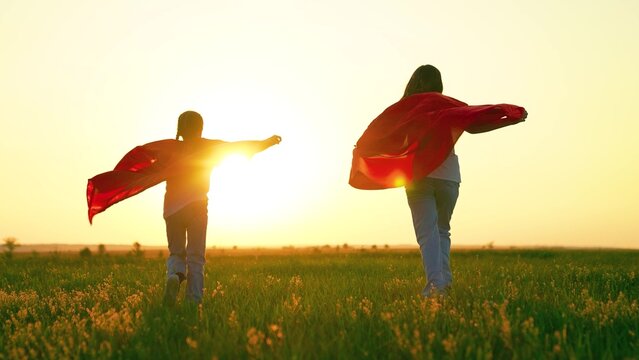 Children heroes in red capes run sunset. Children girls play superheroes in red capes in park on green grass, children dream. Happy children run play superheroes in nature. Kids in capes play heroes - Powered by Adobe