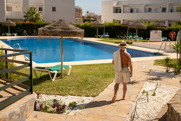 Barefoot elderly man with towel walking to the swimming pool in Spanish resort, enjoying peaceful...