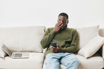 A stressed young Black man in a green sweater sitting on a sofa, using a smartphone while feeling...