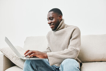 Young Black man smiling while working on a laptop at home in a cozy sweater, capturing a joyful and...