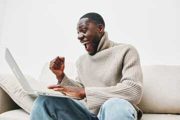 Happy Black man celebrating success on laptop in cozy sweater, expressing joy and excitement while...
