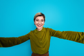 Young woman with short hair in stylish khaki outfit smiling brightly against a blue backdrop,...
