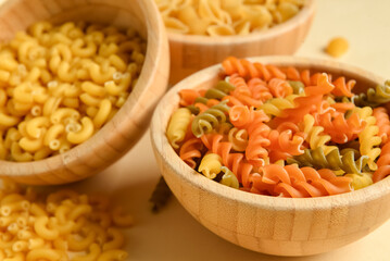 Wooden bowls with raw elbow and fusilli pasta on beige background, closeup