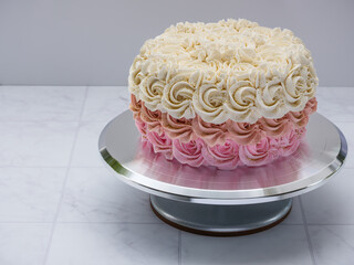 Isolated bakery cake with antique cream and pink rosette swirl pastel ombre frosting and glitter, on a sterling silver raised pedestal platter. Neutral soft gray counter and backdrop.