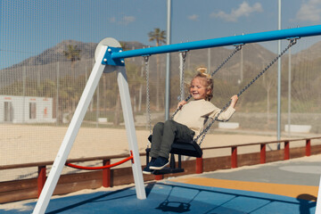 Naklejka premium A young girl swinging happily at the playground, her feet reaching high in the air while enjoying a moment of playful freedom. She gazes ahead with a calm expression