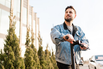 Attractive man in a denim jacket riding electric kick scooter at office buildings background. Driving an ecological mode of transport.