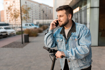 Young and handsome man with electric scooter talk on the phone, standing against the background of office buildings. Using gadgets in work and everyday life.