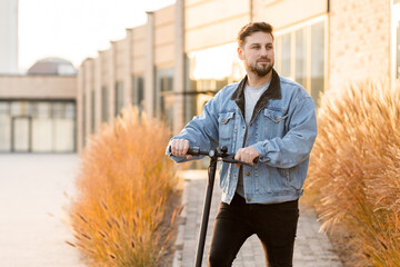 Attractive man in a denim jacket riding electric kick scooter at office buildings background. Driving an ecological mode of transport.