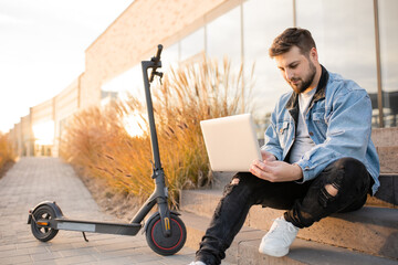 Young and handsome man with a beard sitting on the steps near electric scooter and works at his...