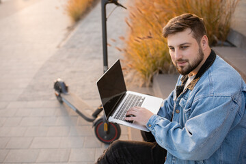 Young and handsome man with a beard sitting on the steps near electric scooter and works at his laptop against the background of office buildings. Freelance work outside the home. 