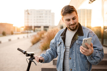 Young and handsome man with electric scooter using smartphone standing against the background of office buildings. Using gadgets in work and everyday life.