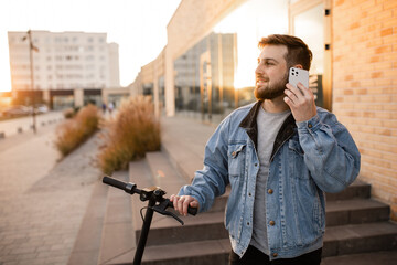 Young and handsome man with electric scooter talk on the phone, standing against the background of office buildings. Using gadgets in work and everyday life.